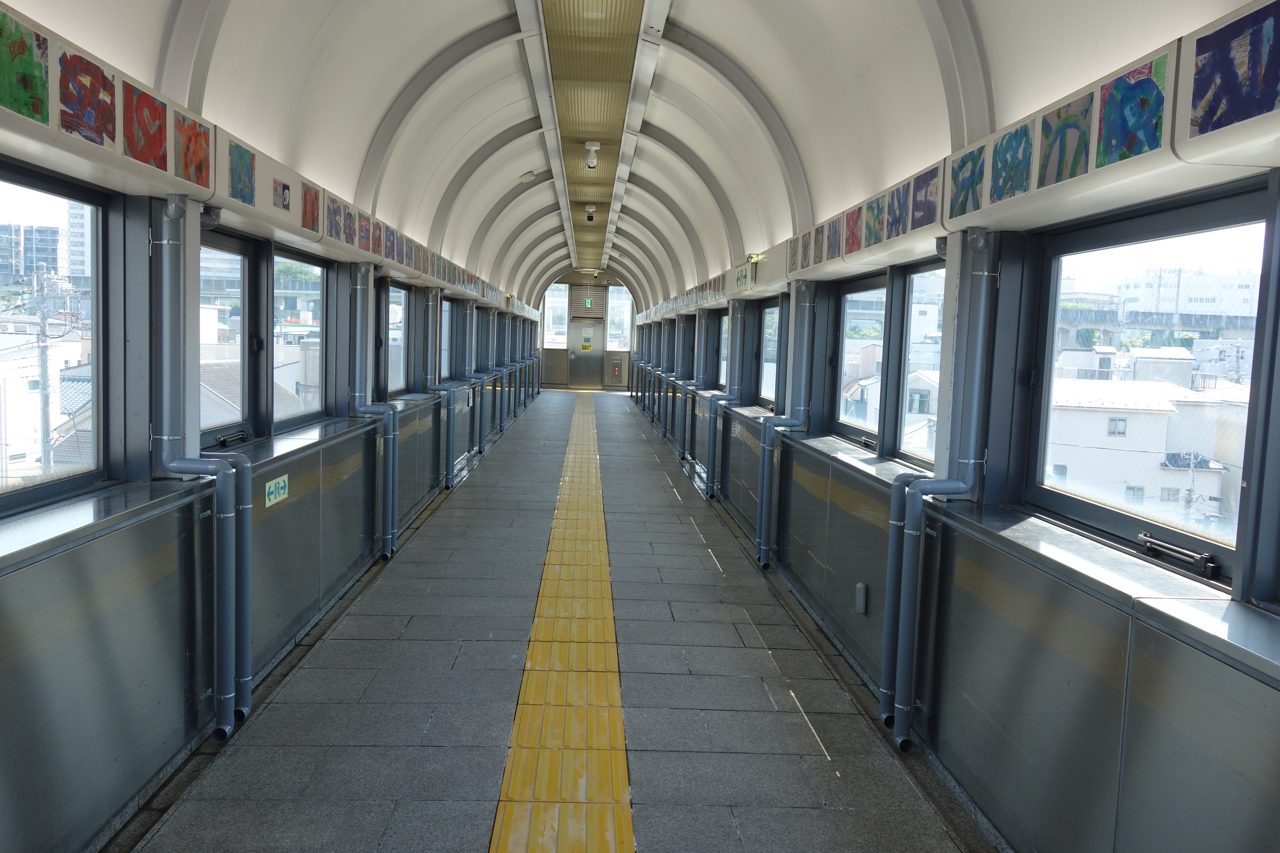 上中里さわやか橋の屋根付き通路。アーチ型の天井と両側のガラス窓、中央の黄色い点字ブロック