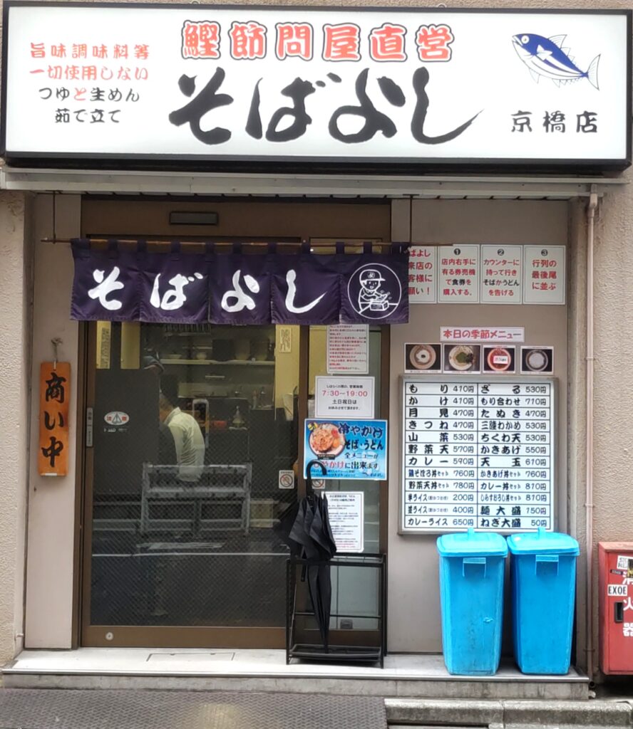 そばよし京橋店の外観。「鰹節問屋直営」の看板と紺色の暖簾、旨味調味料不使用の掲示