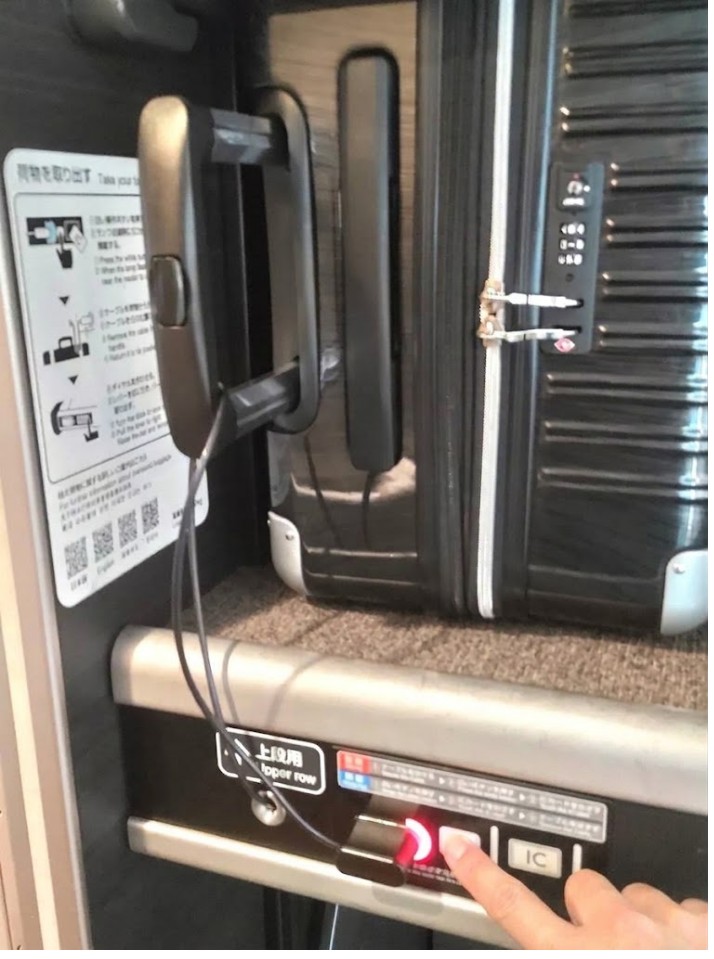 新幹線デッキ荷物置き場の操作パネル。赤ランプ(施錠中)の右にある白いボタンを人差し指で押している操作イメージ(現地写真に手を合成)