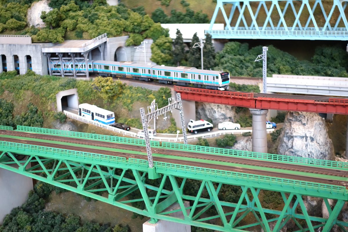 トンネルの脇の線路にいる京浜東北線のE233系。青いトラス橋・赤い橋・緑のトラス橋が重なる渓谷