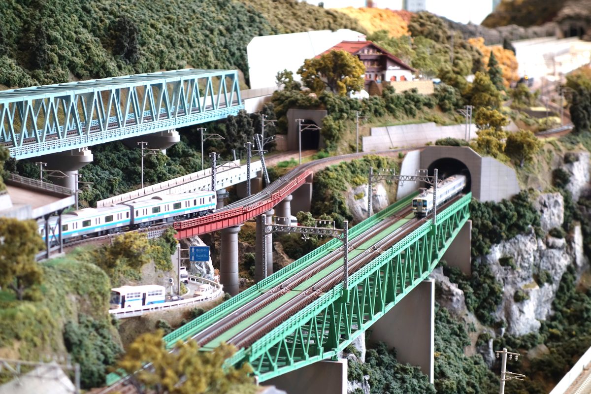 赤い橋の上の京浜東北線のE233系と、緑の鉄橋とトンネルのあいだにいる銀色の電車